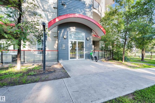 Contemporary building entrance featuring a curved red canopy, grey horizontal siding, and a glass-paneled entry door - #209 10611 117 St Nw, Edmonton, AB - Outdoor