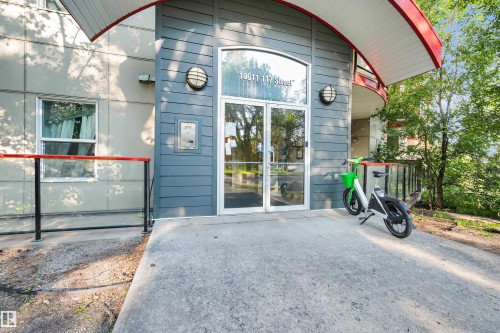 Building entrance featuring a concrete walkway, glass double doors, and a curved red-accented overhang - #209 10611 117 St Nw, Edmonton, AB - Outdoor