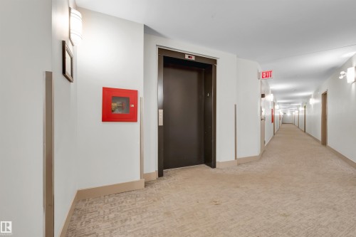Building corridor featuring neutral-toned carpet flooring, wall-mounted light fixtures, white walls, and a dark elevator door - #209 10611 117 St Nw, Edmonton, AB - Indoor Photo Showing Other Room