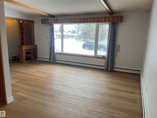 Spacious living area featuring light wood-finish flooring, a large picture window with a wood valance, and a built-in wood shelving unit - 8736 72 Street Nw, Edmonton, AB - Indoor Photo Showing Other Room