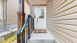 Entryway featuring a white door with decorative glass, beige siding, stone-clad pillar, black metal railing, and concrete steps - 