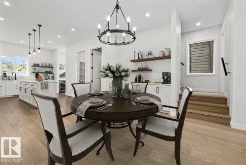 Open-concept main living area featuring wood-finish flooring, a contemporary kitchen with white cabinetry and an island, a built-in beverage station, a dining area with a chandelier, and a stairway with a window - 10934 132 Street, Edmonton, AB - Indoor Photo Showing Dining Room