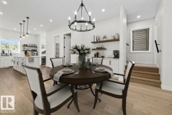 Open-concept main living area featuring wood-finish flooring, a contemporary kitchen with white cabinetry and an island, a built-in beverage station, a dining area with a chandelier, and a stairway with a window - 