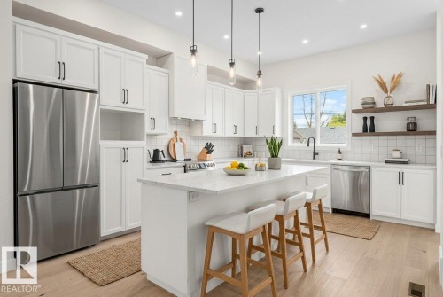 Bright kitchen featuring white shaker cabinetry, stainless steel appliances, a center island with countertop seating, wood-finish flooring, and pendant lighting - 10934 132 Street, Edmonton, AB - Indoor Photo Showing Kitchen With Upgraded Kitchen