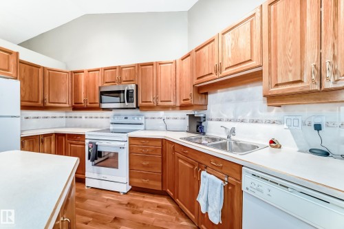 Kitchen featuring wood cabinetry, light-toned countertops, a stainless steel over-the-range microwave, a double basin sink, and wood-finish flooring - 29 929 Picard Drive, Edmonton, AB - Indoor Photo Showing Kitchen With Double Sink