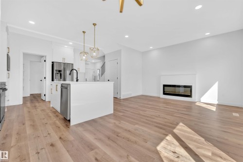 Open-concept living space featuring wood-finish flooring, a contemporary kitchen island with pendant lighting, stainless steel appliances, and a modern fireplace - 523 Kleins Court, Leduc, AB - Indoor With Fireplace