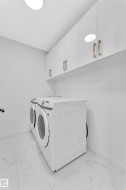 Contemporary laundry space featuring white flat-panel cabinetry with gold-finish hardware, a pair of full-size front-loading machines, and light-toned tile flooring - 