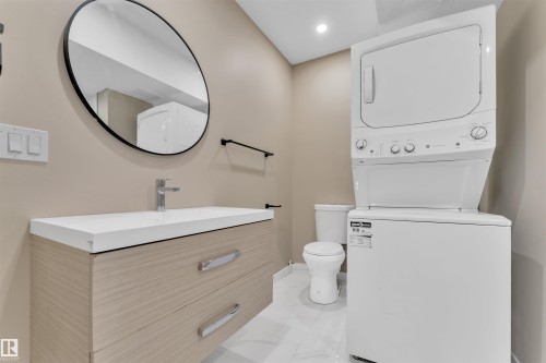 Compact laundry area featuring a stacked washer and dryer, adjacent to a vanity with light wood-finish cabinetry, a white countertop, and a circular wall mirror - 523 Kleins Court, Leduc, AB - Indoor