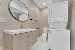 Compact laundry area featuring a stacked washer and dryer, adjacent to a vanity with light wood-finish cabinetry, a white countertop, and a circular wall mirror - 
