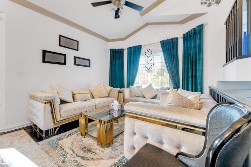 Living area featuring a bay window with teal drapery and sheer curtains, a ceiling fan, and a staircase with dark banister - 7920 7 Avenue, Edmonton, AB 