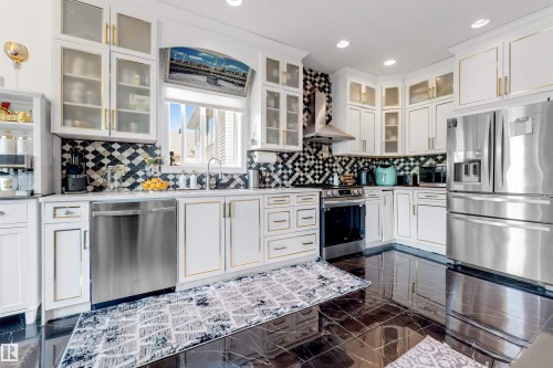 Modern kitchen featuring black marble-look flooring, white cabinetry with gold trim and hardware, a geometric pattern tile backsplash, stainless steel appliances, and recessed lighting - 7920 7 Avenue, Edmonton, AB 