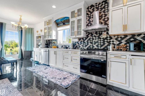 Gourmet kitchen featuring sleek white cabinetry with gold-tone hardware, a striking black and white arabesque tile backsplash, and stainless steel appliances - 7920 7 Avenue, Edmonton, AB 