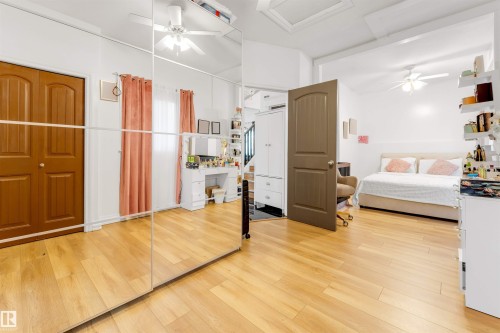 Room featuring wood-finish flooring, full-wall mirrored closet doors, a ceiling fan, and a brown paneled door - 7920 7 Avenue, Edmonton, AB 