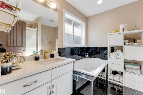 Bathroom featuring a jetted tub, black marble-look tile, white vanity with an integrated sink, gold-finish fixtures, and a frameless mirror - 7920 7 Avenue, Edmonton, AB 