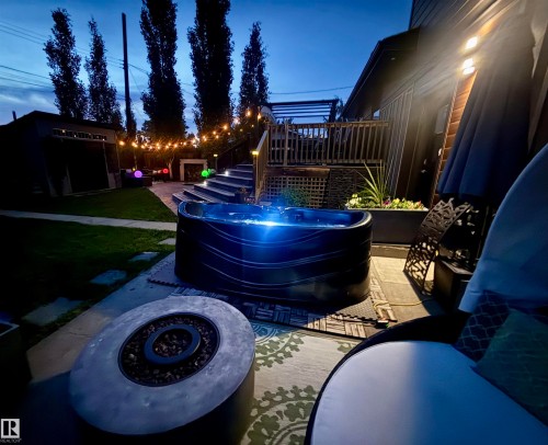 Outdoor hot tub with integrated lighting, a stone fire pit, and string lights illuminating the patio and lawn - 8822 161 Street, Edmonton, AB 