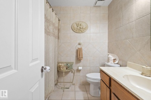 Bathroom with light-colored tile walls, a wood-finish vanity with an integrated sink, a white toilet, and a shower with a patterned curtain - 18126 98 Avenue, Edmonton, AB 