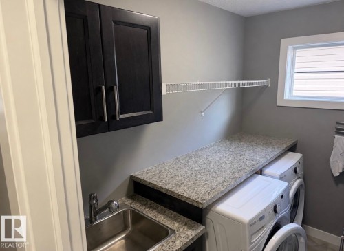 Utility room featuring a stainless steel sink, dark wood-finish cabinetry, light speckled countertops, a white wire shelf, and a window with horizontal blinds - 16505 133 Street, Edmonton, AB 