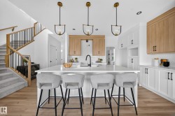 Modern kitchen featuring a large central island with seating, pendant lighting, white cabinetry, wood-finish flooring, and a staircase with wood handrails and black balusters - 