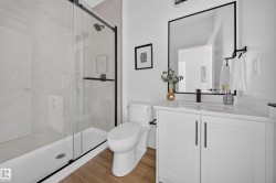 Contemporary bathroom featuring a spacious glass-enclosed shower with black trim, a large white vanity with an integrated sink, black fixtures, and wood-finish flooring - 