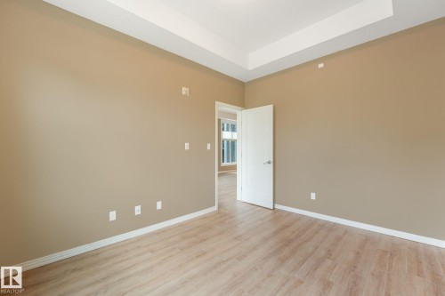 Spacious room featuring a tray ceiling, light wood-finish flooring, neutral wall paint, white trim, and a white interior door with contemporary hardware - 126 10531 117 Street, Edmonton, AB 