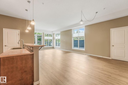 Open-concept living area featuring wood-finish flooring, multiple windows, high ceilings, and a kitchen island with a built-in sink and pendant lighting - 126 10531 117 Street, Edmonton, AB 