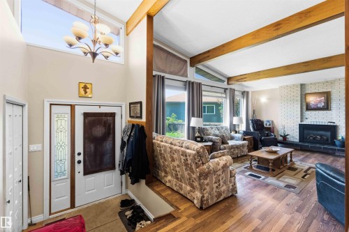 Expansive living area featuring vaulted ceilings with exposed wood beams, a prominent white brick fireplace with a black hearth, and wood-finish flooring - 13121 122 Street, Edmonton, AB 