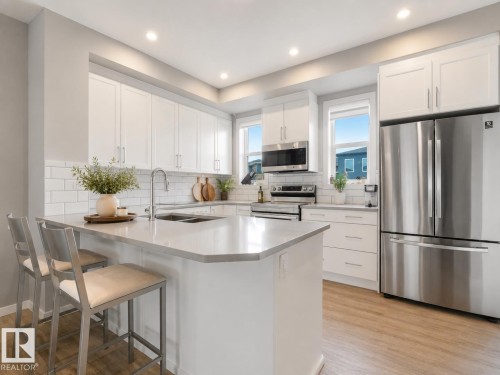 Bright kitchen featuring an expansive center island with seating, white shaker-style cabinetry, stainless steel appliances, subway tile backsplash, and light wood-finish flooring - 516 35 Street, Edmonton, AB 