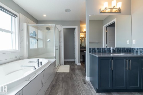 Bathroom featuring a deep soaking tub with tile surround, a glass-enclosed shower, a dark-toned vanity with an integrated sink, a tiled backsplash, and wood-finish flooring - 923 Mcconachie Boulevard, Edmonton, AB 