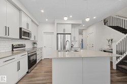 Contemporary kitchen featuring a large island with an undermount sink, white cabinetry with black hardware, stainless steel appliances, and wood-finish flooring - 