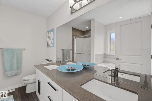 Bathroom featuring a dual vanity with undermount sinks and chrome fixtures, a large wall-mounted mirror, wood-finish flooring, and a window above the soaking tub - 432 Roberts Crescent, Leduc, AB 