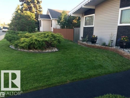 Landscaped front yard featuring a verdant lawn, a stone-edged garden bed with mature shrubs, and a decorative retaining wall - 13304 70 Street, Edmonton, AB 