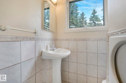 Bathroom featuring light-toned tile wainscoting with decorative border, pedestal sink with chrome faucet, and a window framing mature trees - 
