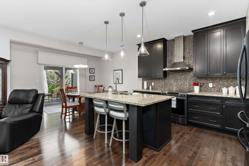 Open concept kitchen featuring dark wood cabinetry, a granite-topped island with seating, a mosaic tile backsplash, stainless steel range hood, and wood-finish flooring - 1097 Armitage Crescent, Edmonton, AB 