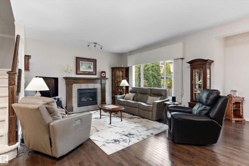 Living area featuring dark wood-finish flooring, a fireplace with a stone surround and wood mantel, a large window with exterior greenery, and a staircase with wood newel posts and carpeted treads - 1097 Armitage Crescent, Edmonton, AB 