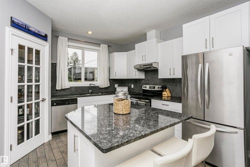 Modern kitchen featuring a central island with a stone countertop, white shaker-style cabinetry, stainless steel appliances, a dark tile backsplash, and wood-finish flooring - 11619 125 Street, Edmonton, AB 