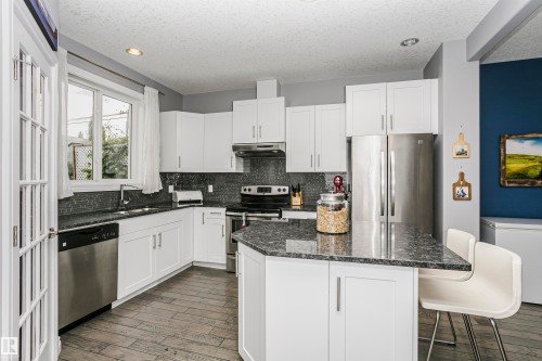 Modern kitchen featuring white shaker cabinetry, stainless steel appliances, dark stone countertops, a mosaic tile backsplash, and wood-finish flooring - 11619 125 Street, Edmonton, AB 