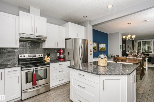 Contemporary kitchen featuring white cabinetry, stainless steel appliances, a dark tile backsplash, and a central island with a granite countertop - 11619 125 Street, Edmonton, AB 