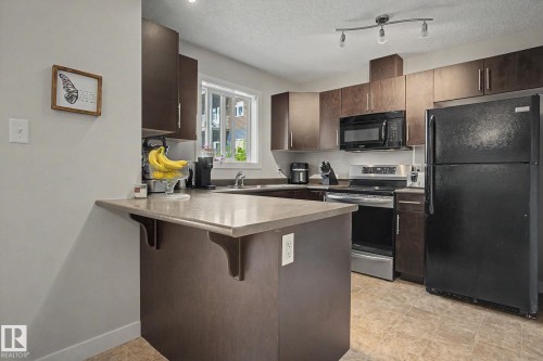 Kitchen featuring dark wood-finish cabinetry, a black refrigerator, stainless steel range and microwave, light-toned countertops, and a track lighting fixture - 25 12004 22 Avenue, Edmonton, AB - Indoor Photo Showing Kitchen