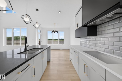 Modern kitchen featuring a large island with an undermount sink and black faucet, light wood-finish flooring, white cabinetry with gold and black hardware, stone countertops, and a subway tile backsplash - 803 30 Avenue, Edmonton, AB 