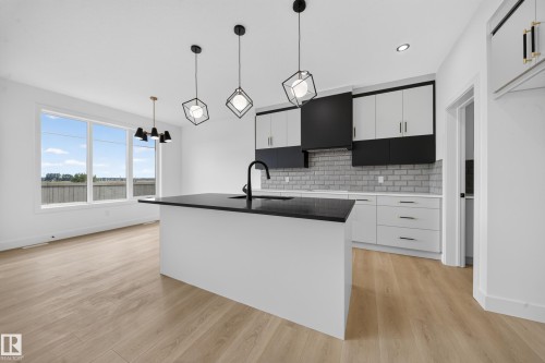 Kitchen featuring a large island with a black sink and faucet, light wood-finish flooring, white cabinetry with black accents, a light gray brick-patterned backsplash, and modern pendant lighting - 803 30 Avenue, Edmonton, AB 