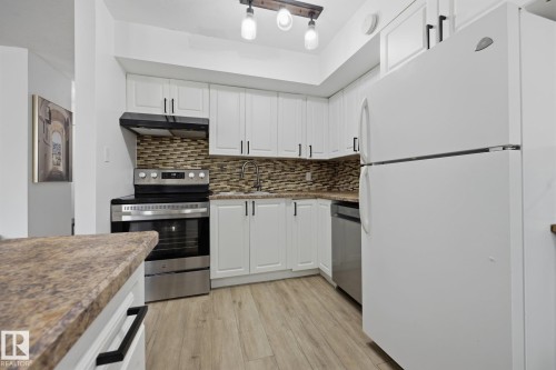 Galley kitchen featuring white cabinetry with matte black hardware, stainless steel appliances, mosaic tile backsplash, wood-finish flooring, and a peninsula with a stone-look countertop - 105 16428 109 Street, Edmonton, AB 