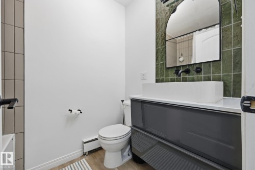 Modern bathroom featuring a rectangular vessel sink, wall-mounted matte black faucet, green square tile backsplash, dark gray vanity with integrated countertop, and a decorative arched mirror - 105 16428 109 Street, Edmonton, AB 