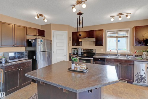 Kitchen featuring a central island with a speckled countertop, dark wood-finish cabinetry, stainless steel appliances, and track lighting fixtures - 1216 37C Avenue Nw, Edmonton, AB - Indoor Photo Showing Kitchen With Double Sink