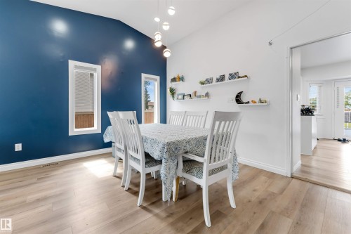 Vaulted ceiling dining area featuring a distinctive blue accent wall, wood-finish flooring, multiple windows with white trim, and a contemporary multi-bulb pendant light fixture - 3332 43 Street, Edmonton, AB 