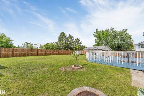 Expansive backyard featuring a large wooden deck with white railing, a manicured lawn, a privacy fence, and a built-in fire pit with paver surround - 3332 43 Street, Edmonton, AB 