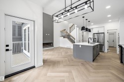 Open-concept living space featuring wood-finish herringbone flooring, a contemporary kitchen island with a white countertop and dark pendant lighting, and a modern linear fireplace - 
