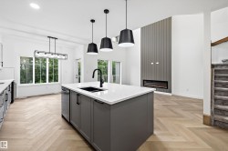 Spacious open-concept living area featuring a kitchen island with stone countertop and integrated sink, chevron wood-finish flooring, a contemporary linear fireplace with a vertical panel accent wall, and large windows providing abundant natural light - 