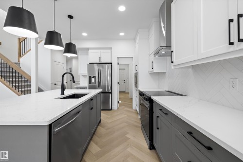 Modern kitchen featuring a large central island with a built-in sink and a matte black faucet, stainless steel appliances, a herringbone tile backsplash, quartz countertops, and wood-finish flooring - 917 Elderberry Landing, Edmonton, AB 