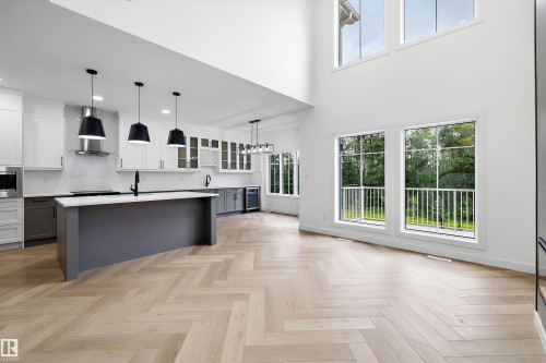 Modern kitchen featuring a large central island, white cabinetry, herringbone wood-finish flooring, and high ceilings with expansive windows - 917 Elderberry Landing, Edmonton, AB 
