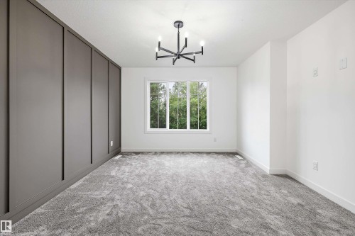 Room featuring a modern black chandelier, a large white-framed window, textured gray carpeting, a paneled accent wall, and clean white walls - 917 Elderberry Landing, Edmonton, AB 